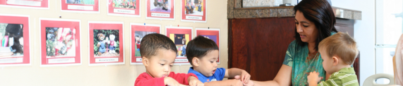 A teacher sitting with three young children, helping them with a craft activity in a classroom with pictures displayed on the wall.