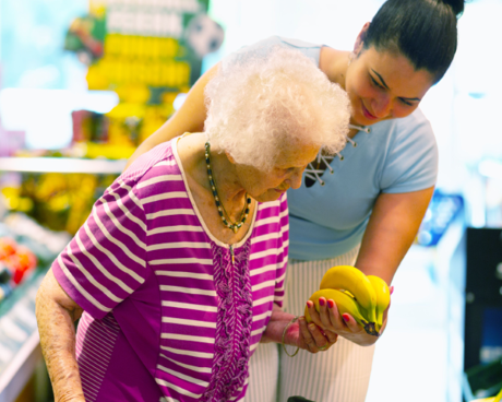 An elderly man wearing a face mask selects fruits and vegetables in a grocery store, while holding a shopping cart.