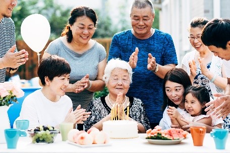 Family gathered around an elderly woman celebrating her birthday outdoors, with a cake and decorations on the table.