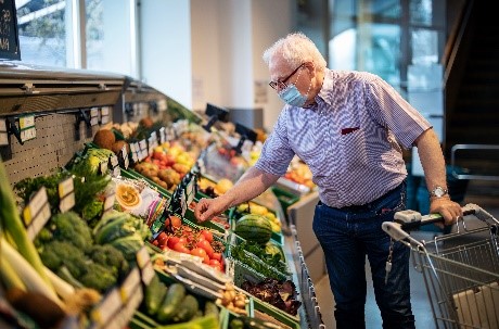 An elderly man wearing a face mask selects fruits and vegetables in a grocery store, while holding a shopping cart.