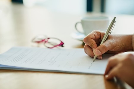 Person writing on a paper at a table with a pen; glasses and a coffee cup are in the background.