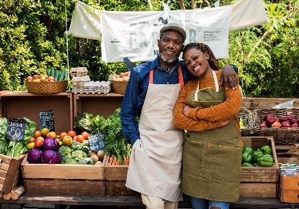 A man and woman stand side by side at a produce stand, surrounded by a variety of fresh fruits and vegetables.