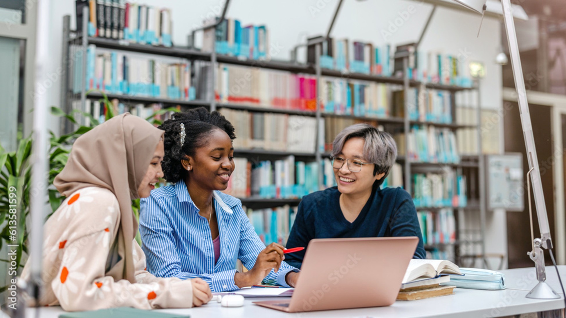 Three diverse students smiling and collaborating at a table in a library with bookshelves in the background.